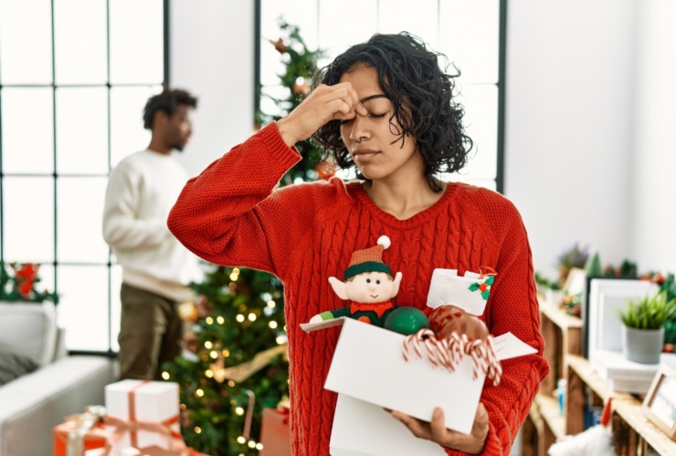 A woman in a holiday sweater, holding gifts, in front of a Christmas tree, holds her hand up to her forehead to show that she is stressed.
