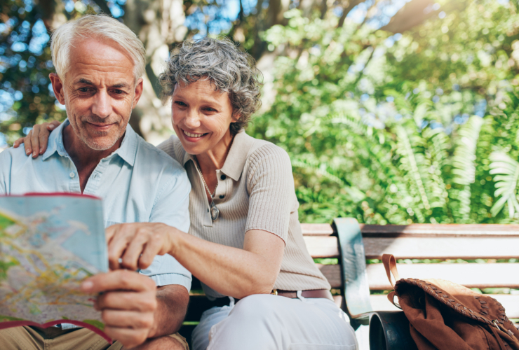Portrait of happy man and woman reading map while sitting on a park bench. Senior couple travel tips.